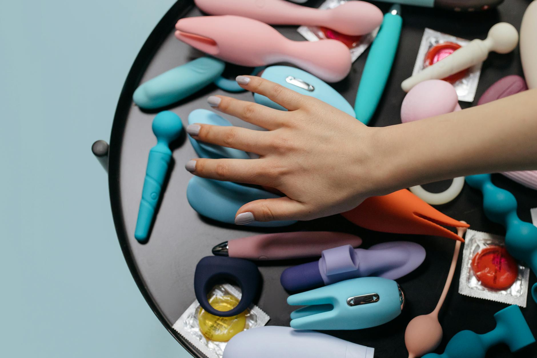 Hands holding and exploring a variety of colorful intimate wellness toys on a table.
