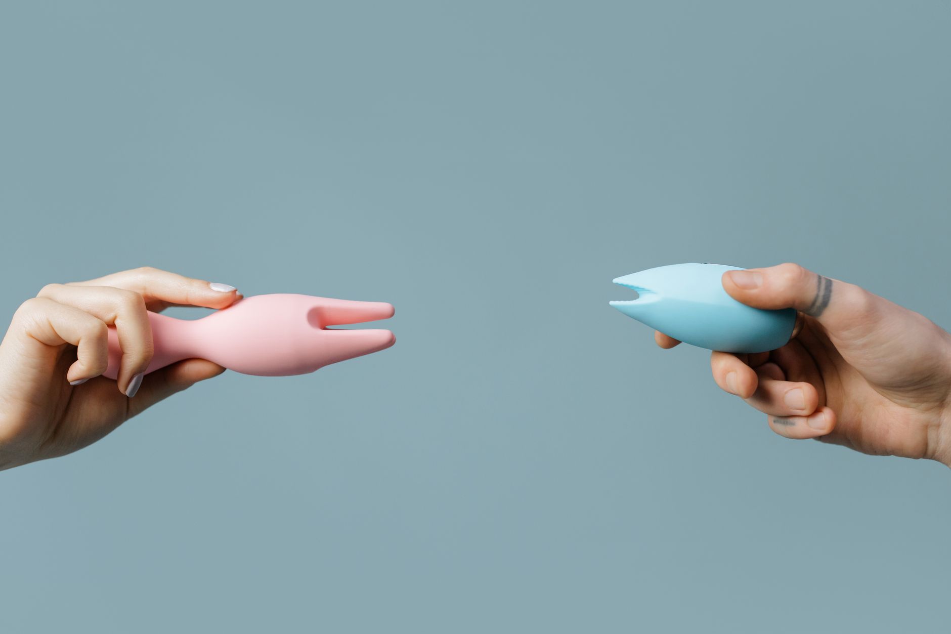 Two hands holding silicone vibrators against a pastel background